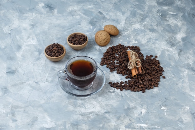 Kaffeebohnen mit hohem Blickwinkel in Schalen mit Tasse Kaffee, Kekse, Zimt auf hellblauem Marmorhintergrund. horizontal