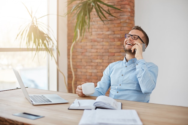 Kaffeepause bei der Arbeit. Erwachsener rasierter Geschäftsmann in den Gläsern, die im bequemen Büro sitzen