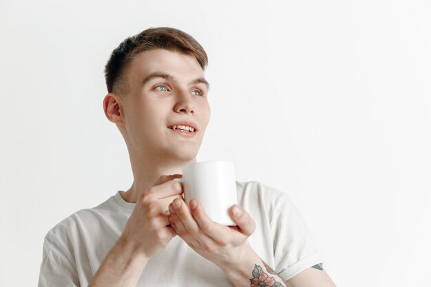 Kaffeepause machen. Hübscher junger Mann, der Kaffeetasse hält, lächelnd, während gegen grauen Studiohintergrund steht