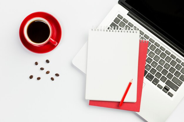 Kaffeetasse, Kaffeebohnen mit gewundenem Notizblock auf einem offenen Laptop gegen weißen Hintergrund