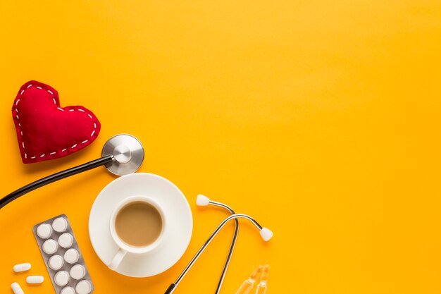Kaffeetasse; Tabletten in Blisterpackungen; Stethoskop und genähte Herzform vor gelbem Hintergrund