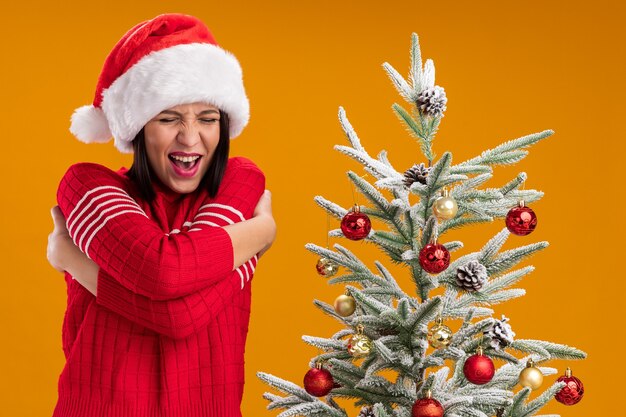 Kaltes junges Mädchen, das Weihnachtsmütze trägt, die nahe geschmücktem Weihnachtsbaum steht, der sich mit fest geschlossenen Augen auf orange Wand lokalisiert umarmt