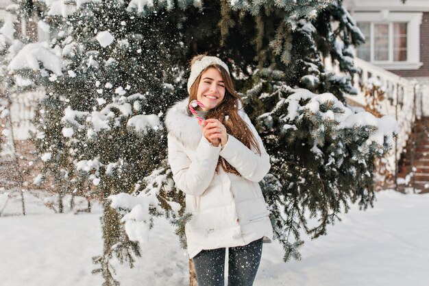 Kaltes Winterwetter in der Stadt und lustiges tolles genossenes Mädchen mit Lollypop auf Fri-Baum-Hintergrund. Warme Kleidung, fröhliche Stimmung, fallender Schnee, positive Emotionen, neues Jahr, süße gefrorene Winterzeit