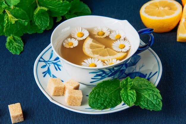 Kamillentee in einer Tasse und Zitronenscheiben, braunen Zuckerwürfeln und grünen Blättern Seitenansicht auf einem dunklen Tischset-Hintergrund