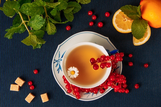 Kamillentee mit roten Johannisbeeren, Zitronen, Zuckerwürfeln und Blättern in einer Tasse und Sauce auf dunklem Tischset-Hintergrund, flach liegen.