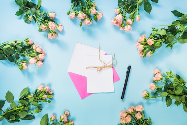 Karte; Stift und rosa Umschlag umgeben mit Strauß rosa Rosen vor blauem Hintergrund