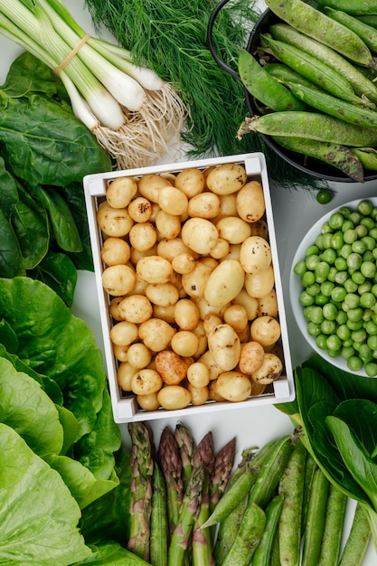Kartoffeln in einer Holzkiste mit grünen Hülsen, Erbsen, Dill, Frühlingszwiebeln, Spinat, Sauerampfer, Salat, Spargel Draufsicht auf einer weißen Wand