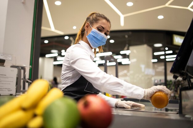 Kassiererin im Supermarkt, die hygienische Schutzmaske und Handschuhe trägt, während riskanter Job wegen Koronavirus-Pandemie arbeitet