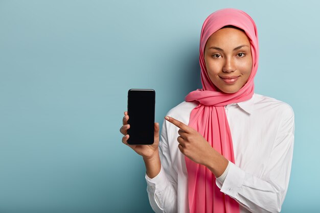 Kaufen Sie dieses Gerät! Erfreute dunkelhäutige Frau im rosa Schleier, zeigt auf Smartphone