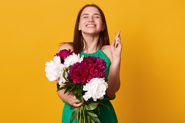 Kaukasierfrau hebt die Daumen, betet um Glück, hat einen erfreuten und glücklichen Ausdruck, trägt grünes Sommerkleid, hält weiße und burgunderfarbene Pfingstrosen, glaubt an Glück.