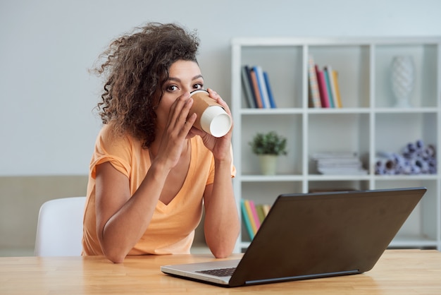 Kaukasische frau, die bei tisch zu hause mit laptop sitzt und mitnehmerkaffee trinkt