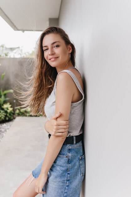 kaukasische Frau im Jeansrock, der mit inspiriertem Gesichtsausdruck schaut. Außenaufnahme des hübschen braunhaarigen Mädchens, das nahe Wand steht und ihren Arm berührt.