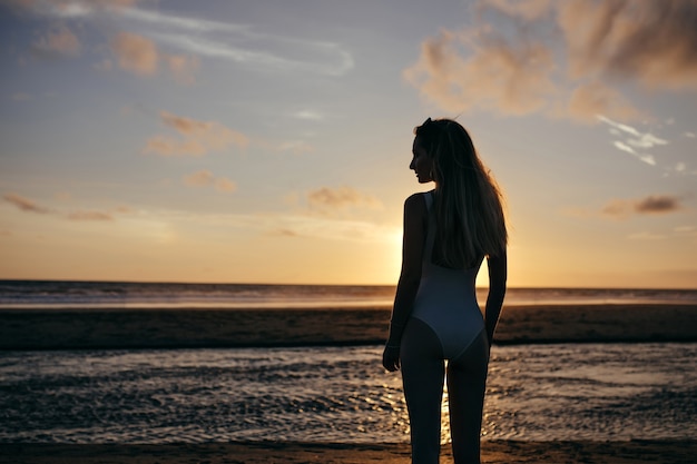 kaukasische Frau trägt weißen Badeanzug im Urlaub. Sorglose junge Dame, die Abend am Meer genießt und schönen Sonnenuntergang betrachtet.