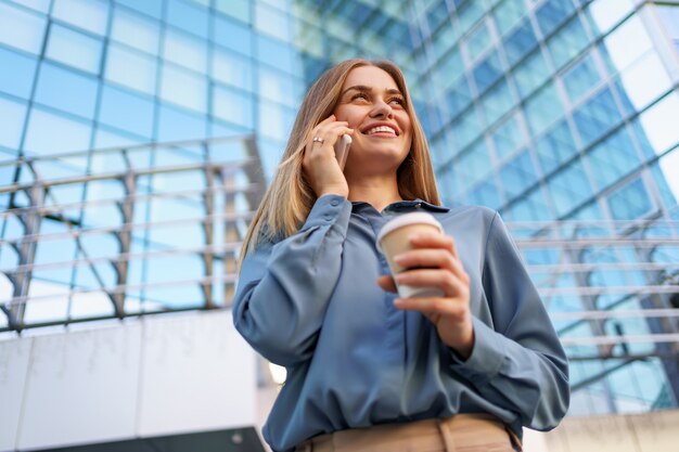 Kaukasische Geschäftsfrau, die telefonisch Kaffee hält, um zu gehen. Eine erfolgreiche europäische Frau, die am Telefon spricht und auf einem modernen Bürogebäude steht