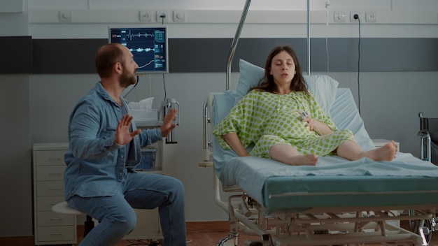 Kaukasische schwangere frau, die schmerzen durch kontraktionen hat, während sie im krankenbett sitzt. junge person, die sich auf die geburt eines kindes bei der mutterschaft vorbereitet, händchen haltend mit dem vater des babys
