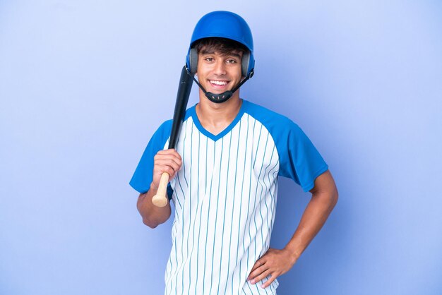 Kaukasischer baseballspieler mit helm und schläger isoliert auf blauem hintergrund, der mit den armen an der hüfte posiert und lächelt