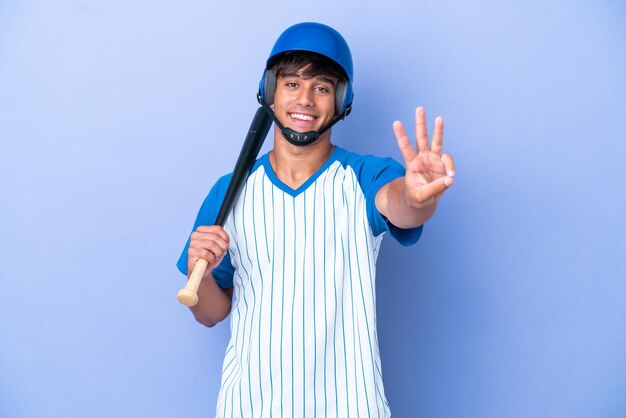 Kaukasischer baseballspieler mit helm und schläger isoliert auf blauem hintergrund glücklich und zählt drei mit den fingern