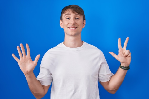 Kostenloses Foto kaukasischer blonder mann, der über blauem hintergrund steht und mit den fingern nummer acht nach oben zeigt, während er selbstbewusst und glücklich lächelt.