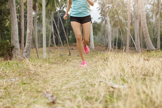 Kaukasischer Frauenjogger mit schönem passendem Körper, der auf Gras im tropischen Wald läuft. Junger weiblicher Läufer, der blaues Sportoberteil und schwarze Shorts trägt, die draußen an sonnigem Tag trainieren.