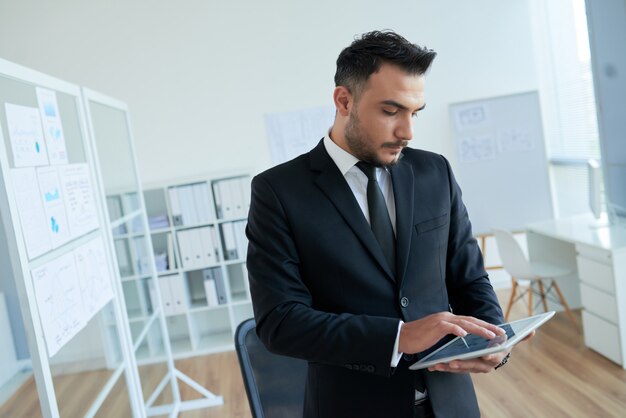 Kaukasischer Geschäftsmann in der intelligenten Klage, die im Büro steht und Tablette verwendet