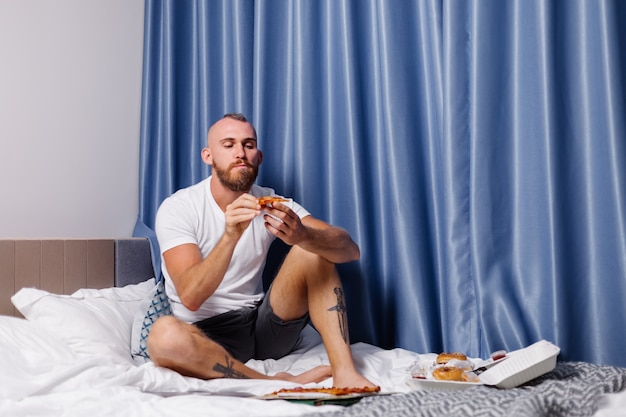 Kostenloses Foto kaukasischer mann, der fastfood zu hause im schlafzimmer auf bett hat