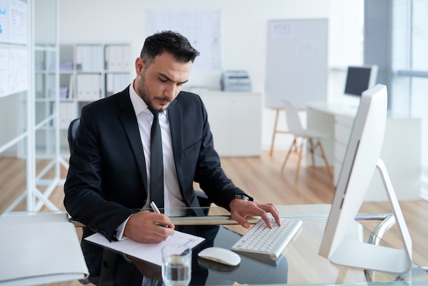 Kaukasischer Mann im Anzug, der im Büro sitzt, an Computer arbeitet und auf Papier schreibt