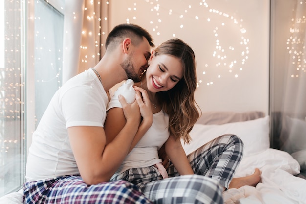Kaukasischer Mann im karierten Pyjama, der seine Frau küsst. Lächelnde blonde Frau in Nachtwäsche, die mit Freund auf Bett sitzt.