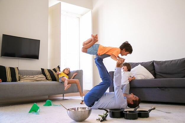 Kaukasischer Vater, der Sohn auf Beinen hält und auf Teppich liegt. Glücklicher süßer Junge, der im Wohnzimmer mit Hilfe des Vaters fliegt. Nette Kinder, die zusammen nahe Schüssel und Pfannen spielen. Kindheits- und Wochenendkonzept