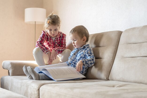 Kaukasisches 6-jähriges Mädchen mit ihrem 4-jährigen Bruder, der zu Hause ein Buch auf der Couch liest