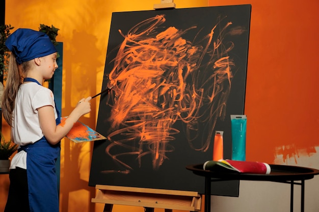 Kaukasisches Kind, das künstlerisches Hobby mit orangefarbener Farbe und Pinsel praktiziert, um ein Meisterwerk auf Leinwand zu schaffen. Künstler mit kreativer Vision und Fähigkeiten bei der Gestaltung von Kunstwerken mit Aquarellfarben.