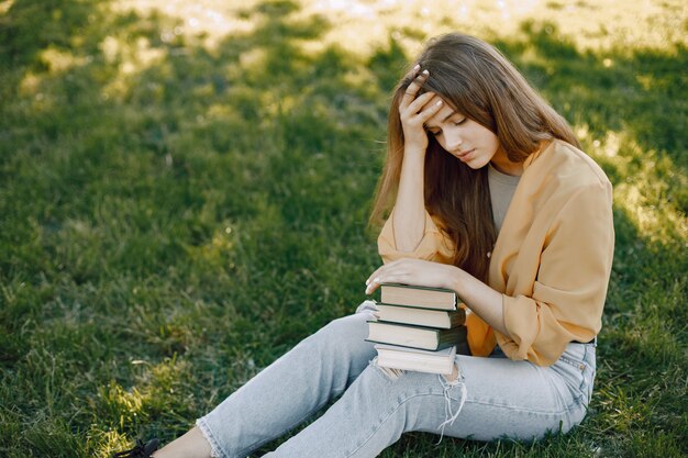 Kaukasisches Mädchen sitzt auf einem Gras in einem Park mit Büchern auf den Knien. Mädchen hält sich die Hand auf die Stirn