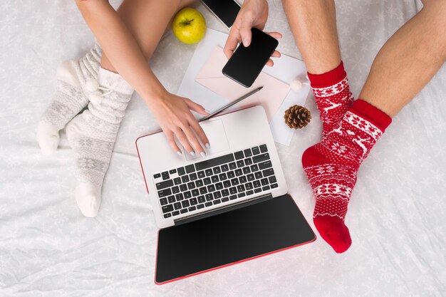 Kaukasisches Paar zu Hause mit Internet-Technologie. Laptop und Telefon für Leute, die auf dem Boden an farbigen Socken sitzen. Weihnachten, Liebe, Lifestyle-Konzept