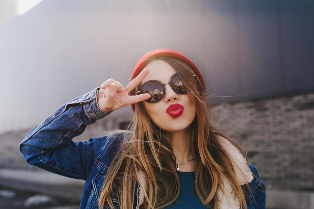 Kaukasisches schönes Mädchen mit roten Lippen, das mit küssendem Gesichtsausdruck posiert Outdoor-Foto einer attraktiven braunhaarigen Dame trägt eine stilvolle Jeansjacke, die ein Friedenszeichen macht