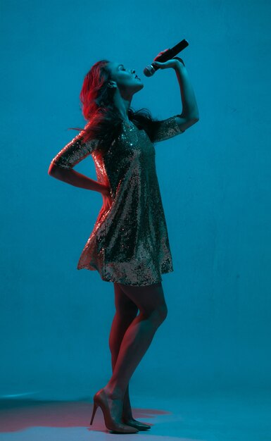 Kaukasisches weibliches Sängerporträt lokalisiert auf blauem Studiohintergrund im Neonlicht. Schönes weibliches Modell im hellen Kleid mit Mikrofon. Konzept menschlicher Emotionen, Gesichtsausdruck, Werbung, Musik, Kunst.