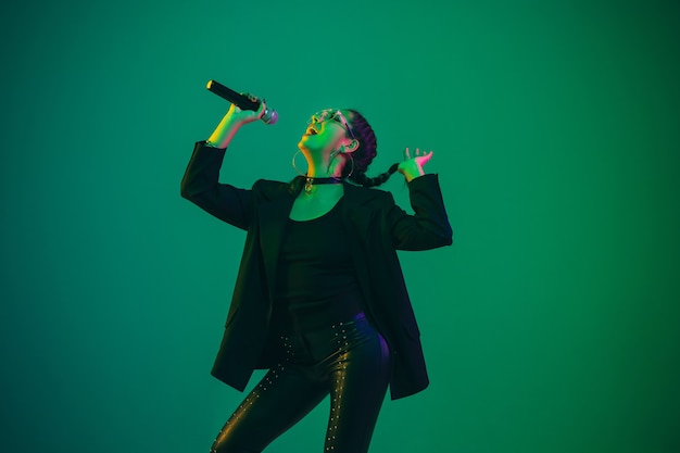 Kaukasisches weibliches Sängerporträt lokalisiert auf grüner Wand im Neonlicht. Schönes weibliches Modell in schwarzer Kleidung mit Mikrofon.