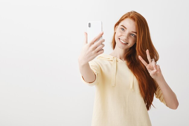 Kawaii rothaariges Mädchen, das selfie mit Friedenszeichen auf Smartphone nimmt