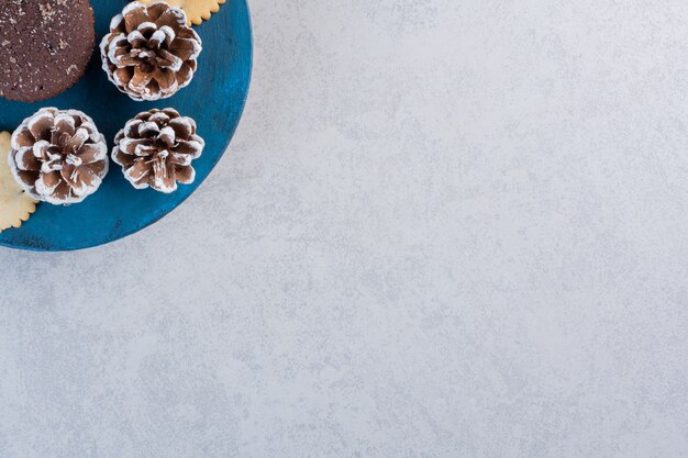 Kekse und ein Schokoladenkuchen auf einem blauen Brett mit Tannenzapfen auf Marmortisch.