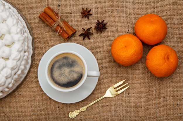 Kekskuchen verziert mit schlagsahne, tasse kaffee, gabel, orangen, sternanis und zimt auf dem tisch mit sacktuch. ansicht von oben