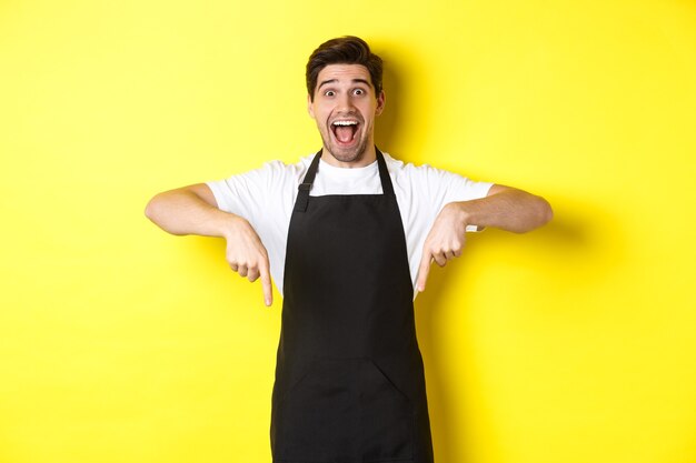 Kellner, Café-Barista in schwarzer Schürze, die mit den Fingern nach unten zeigt, erstaunt aussieht und auf gelbem Hintergrund steht.