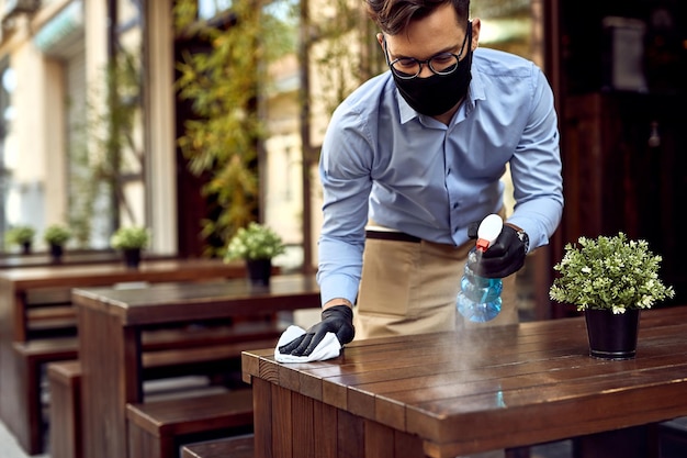 Kellner, der eine schützende Gesichtsmaske trägt, während er Tische im Café im Freien desinfiziert