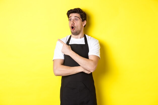 Kellner in schwarzer Schürze, der das Promo-Angebot überprüft, mit dem Finger zeigt und nach links auf Ihr Logo schaut, über gelbem Hintergrund stehend