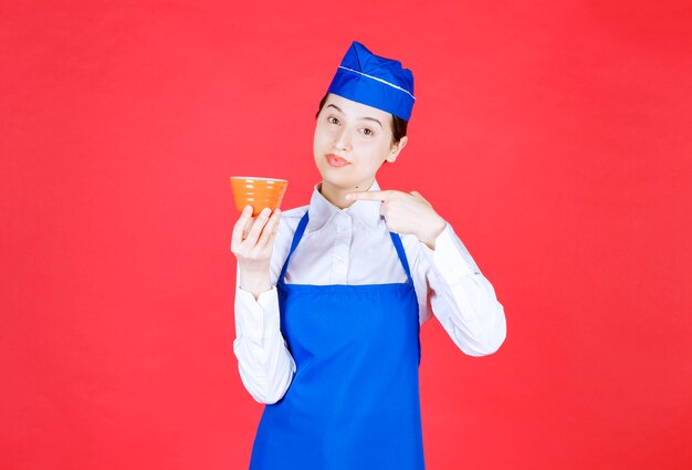 Kellnerin in Uniform, die auf eine orangefarbene Schüssel an der roten Wand zeigt.