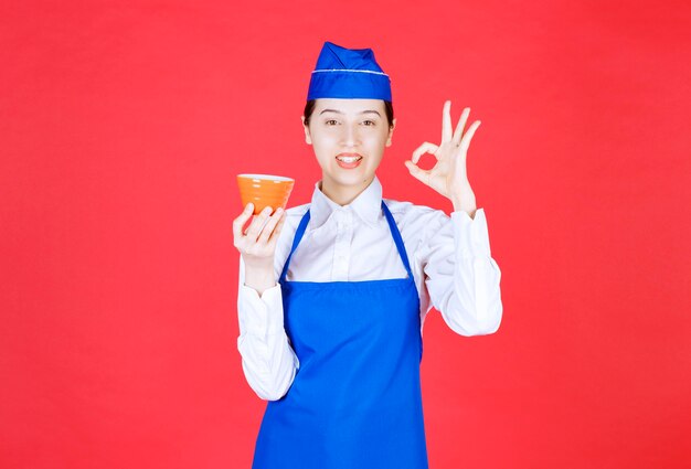 Kellnerin in Uniform, die eine orangefarbene Schüssel hält und eine ok Geste zeigt.