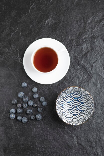 Keramikschale mit köstlichen frischen Blaubeeren und einer Tasse Tee auf schwarzer Oberfläche