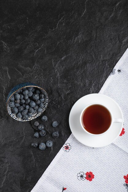 Keramikschale mit köstlichen frischen Blaubeeren und einer Tasse Tee auf schwarzer Oberfläche