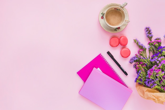Keramische Kaffeetasse; Makronen; Stift; Notizbuch- und Blumenblumenstrauß auf rosa Hintergrund