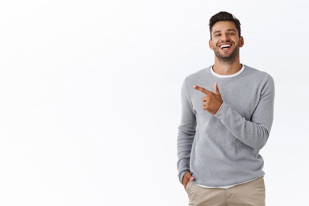 Kerl, der mit einem Freund von der Universität spricht, erinnert sich an lustige, schöne Erinnerungen, die breit lachen und lächeln Attraktives, athletisches, bärtiges männliches Modell im trendigen Pullover, der nach links zeigt und mit stolzem Gesicht kichert