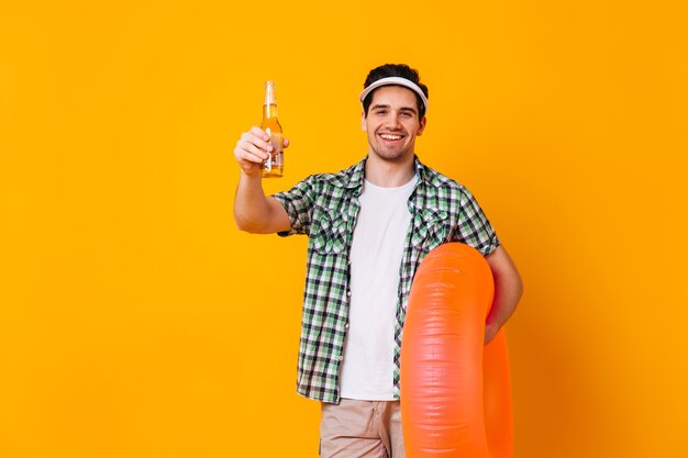 Kerl in der Kappe und im weißen T-Shirt, das Flasche Bier und orange aufblasbaren Kreis auf isoliertem Raum hält.