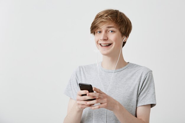 Kerl mit blondem Haar, graues T-Shirt tragend modernes Smartphone über Hochgeschwindigkeits-Internetverbindung, SMS an seine Freunde, breit lächelnd. Moderne Technologie und Kommunikation.