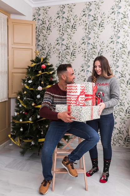 Kerl mit Haufen von Geschenken auf Stuhl und attraktive Dame in der Nähe von Weihnachtsbaum